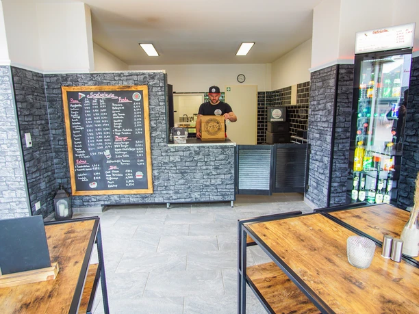 Innenraum von Speedy's Heller Gastraum mit modernen Holztischen, einer Getränkekühlvitrine und einer großen Menütafel an der Wand. Hinter der Theke steht ein Mitarbeiter mit einer Pizzaschachtel. Die Einrichtung wirkt klar strukturiert, barrierefrei zugänglich und auf Selbstbedienung ausgelegt.