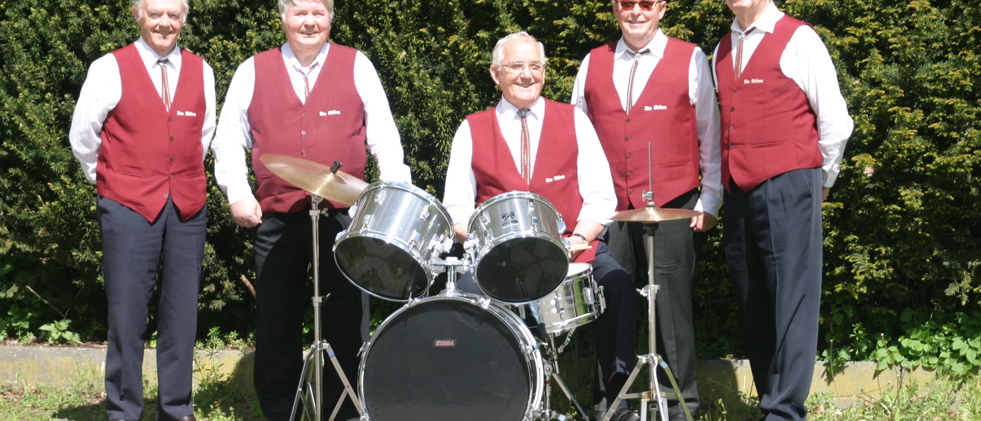 Fünf ältere Herren in roten Westen stehen lächelnd neben einem Schlagzeug, vor grünen Büschen.