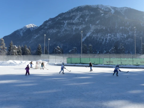 Eisplatz Farchant Eishockeyspielende Kinder, im Hintergrund Zaun, Flutlichtlampen und der schneebedeckte Hohe Fricken