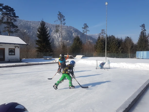 Eisplatz Farchant 2 Eishockeyspielende Kinder mit Tor, im Hintergrund Bäume und der Umkleideraum