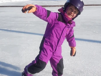 Eisplatz Farchant kleines Mädchen im lila Schneeanzug und lila Helm auf Schlittschuhen auf dem Eisplatz, im Hintergrund Zaun und Bäume