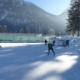 Eisplatz Farchant 5 Personen auf dem im Schatten liegendem Eisplatz. Im Hintergrund das Tor, Zaun, Bäume, Flutlichtlampen sowie der Hohe Fricken