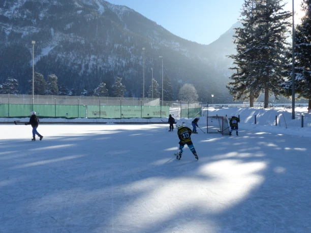 Eisplatz Farchant 5 Personen auf dem im Schatten liegendem Eisplatz. Im Hintergrund das Tor, Zaun, Bäume, Flutlichtlampen sowie der Hohe Fricken
