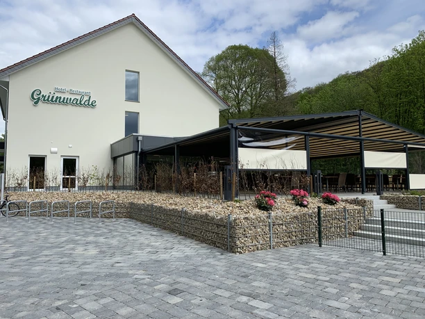 Ein modernes Restaurant mit Außenterrasse, umgeben von grüner Natur und einem gepflasterten Vorplatz.