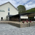 Halle-Aussenansicht-TeutoburgerWald-HotelRestaurantGrünwalde.jpg Ein modernes Restaurant mit Außenterrasse, umgeben von grüner Natur und einem gepflasterten Vorplatz.