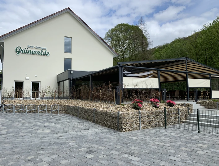Halle-Aussenansicht-TeutoburgerWald-HotelRestaurantGrünwalde.jpg Ein modernes Restaurant mit Außenterrasse, umgeben von grüner Natur und einem gepflasterten Vorplatz.