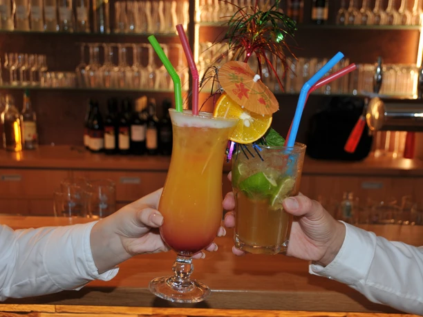 Zwei Cocktails auf einer Theke werden von Menschen in weißen Hemden angestoßen, Hintergrund Flaschen.