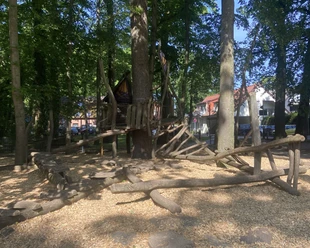 Spielplatz an der Grundschule Spielstruktur aus Holz in einem schattigen Waldabschnitt, umgeben von Bäumen und weichem Bodenbelag.