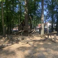 Spielplatz an der Grundschule Spielstruktur aus Holz in einem schattigen Waldabschnitt, umgeben von Bäumen und weichem Bodenbelag.