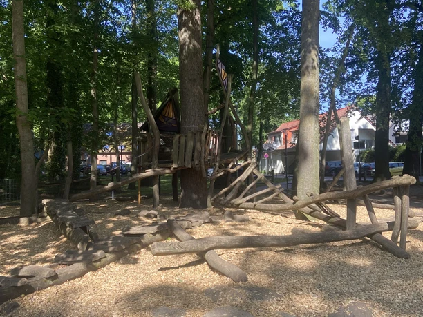 Spielplatz an der Grundschule Spielstruktur aus Holz in einem schattigen Waldabschnitt, umgeben von Bäumen und weichem Bodenbelag.