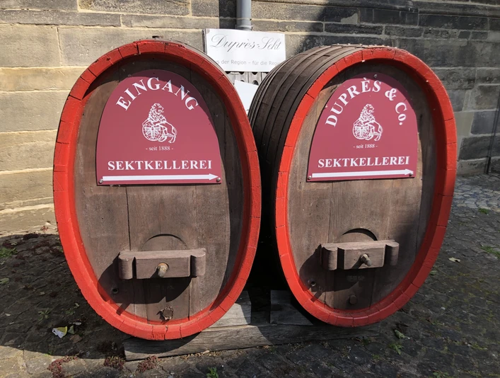 Zwei große Holzfässer mit roten Schildern kennzeichnen den Eingang der Sektkellerei Duprès.Two large wooden barrels with red signs mark the entrance to the Duprès sparkling wine cellar.To store træfade med røde skilte markerer indgangen til Duprès' mousserende vinkælder.Twee grote houten vaten met rode tekens markeren de ingang van de mousserende wijnkelder van Duprès.