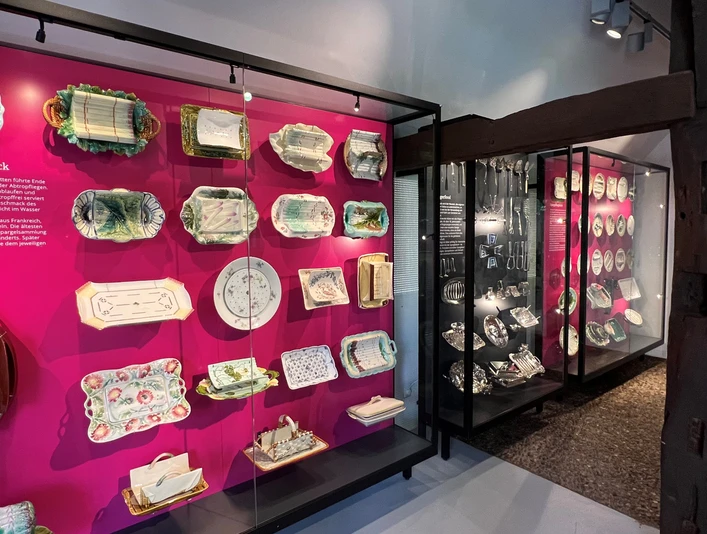 Spargelmuseum Vitrinen mit historischen Spargeltellern und Besteck setzen farbliche Akzente auf magentafarbenem Hintergrund.Display cases with historical asparagus plates and cutlery set colorful accents on a magenta background.Vitrineskabe med historiske aspargestallerkener og bestik sætter farverige accenter på en magenta baggrund.Vitrinekasten met historische aspergeborden en bestek zetten kleurrijke accenten op een magenta achtergrond.