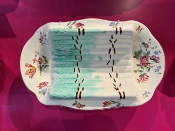 Ein kunstvoll bemalter Porzellanteller ausgestellt auf einem kräftig pinkfarbenen Hintergrund.