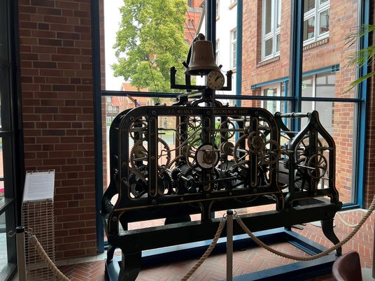 Uhrwerk Historisches Uhrwerk mit Zahnrädern, umgeben von zwei Ziegelwänden und großen Fenstern.