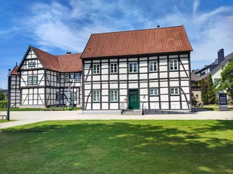 Hoevelhof-Tourist_Information_02-CC_BY_SA-Tourist_Information_Hoevelhof.jpg Fachwerkhaus mit rotem Ziegeldach vor blauem Himmel, umgeben von Grünflächen auf einem sonnigen Tag.
