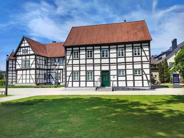 Hoevelhof-Tourist_Information_02-CC_BY_SA-Tourist_Information_Hoevelhof.jpg Fachwerkhaus mit rotem Ziegeldach vor blauem Himmel, umgeben von Grünflächen auf einem sonnigen Tag.