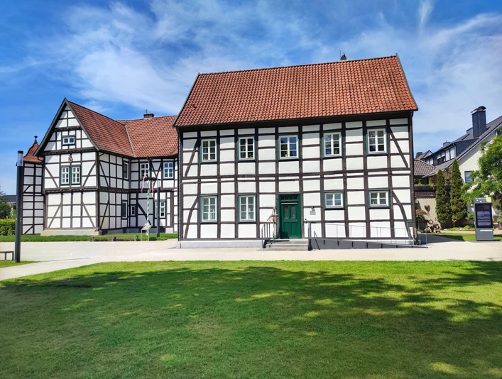 Hoevelhof-Tourist_Information_02-CC_BY_SA-Tourist_Information_Hoevelhof.jpg Fachwerkhaus mit rotem Ziegeldach vor blauem Himmel, umgeben von Grünflächen auf einem sonnigen Tag.