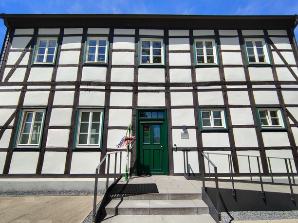 Fachwerkgebäude mit grüner Eingangstür, weißen Wänden und dunklen Holzbalken unter blauem Himmel.
