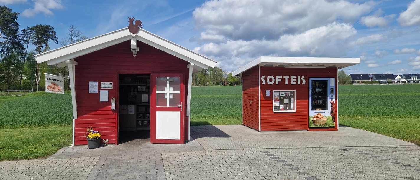 Zwei rote Holzhütten in ländlicher Umgebung, eine beschriftet mit "SOFTEIS", flankiert von Feldern.