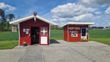 Hof Bröker Verkaufshütten.jpg Zwei rote Holzhütten in ländlicher Umgebung, eine beschriftet mit "SOFTEIS", flankiert von Feldern.