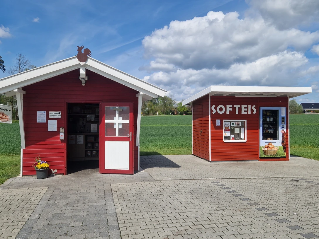 Hof Bröker Verkaufshütten.jpg Zwei rote Holzhütten in ländlicher Umgebung, eine beschriftet mit "SOFTEIS", flankiert von Feldern.