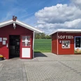 Hof Bröker Verkaufshütten.jpg Zwei rote Holzhütten in ländlicher Umgebung, eine beschriftet mit "SOFTEIS", flankiert von Feldern.