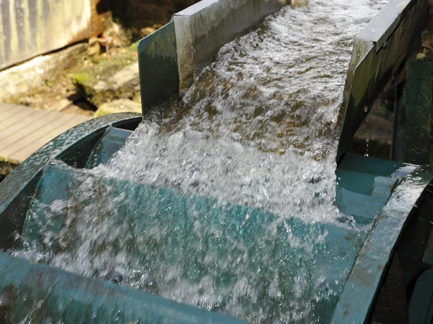 Wasserrad am Oelchenshammer Wasser fließt dynamisch über ein rustikales Wasserrad, umgeben von natürlicher Vegetation.