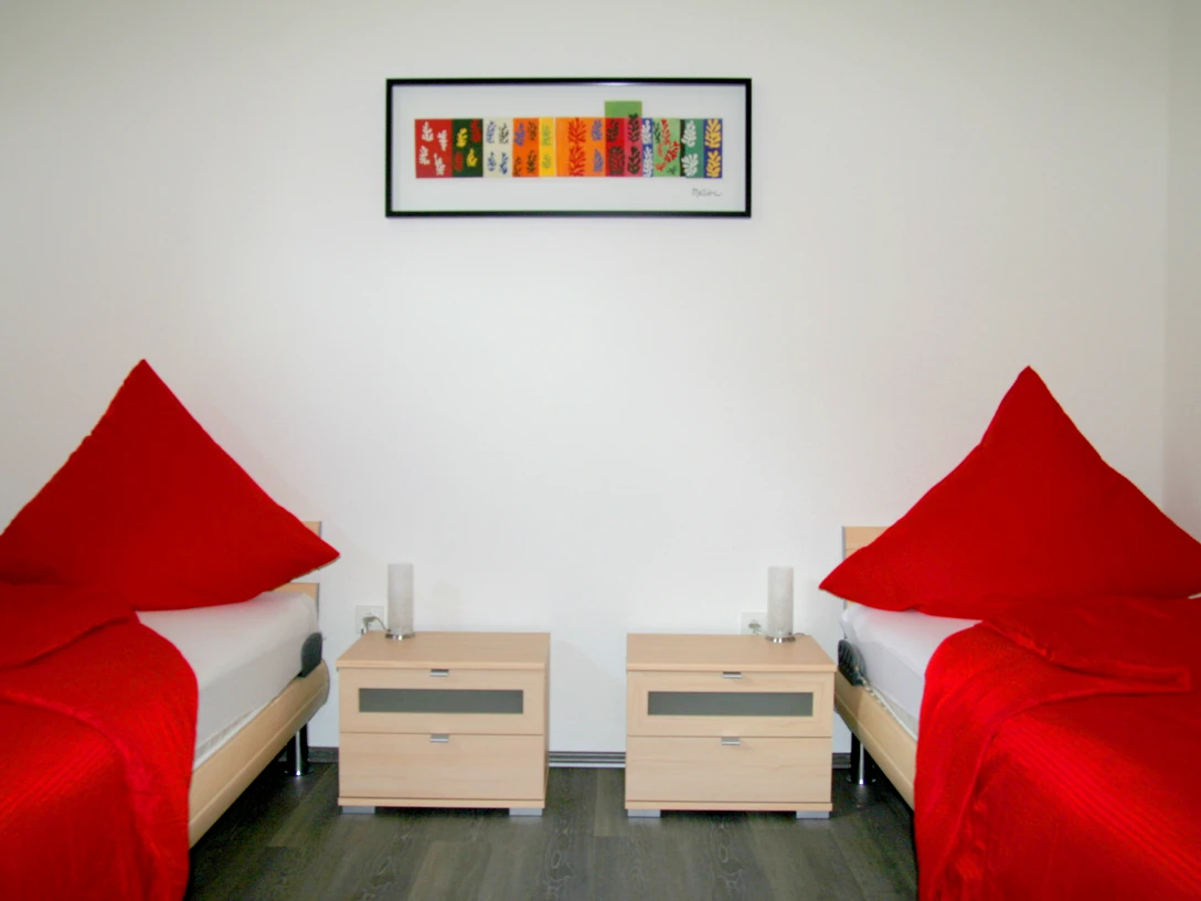 appartmentshaus_senne.jpg Zwei Einzelbetten mit roten Tagesdecken und zweckmäßigen Nachttischen in einem hellen Raum.
