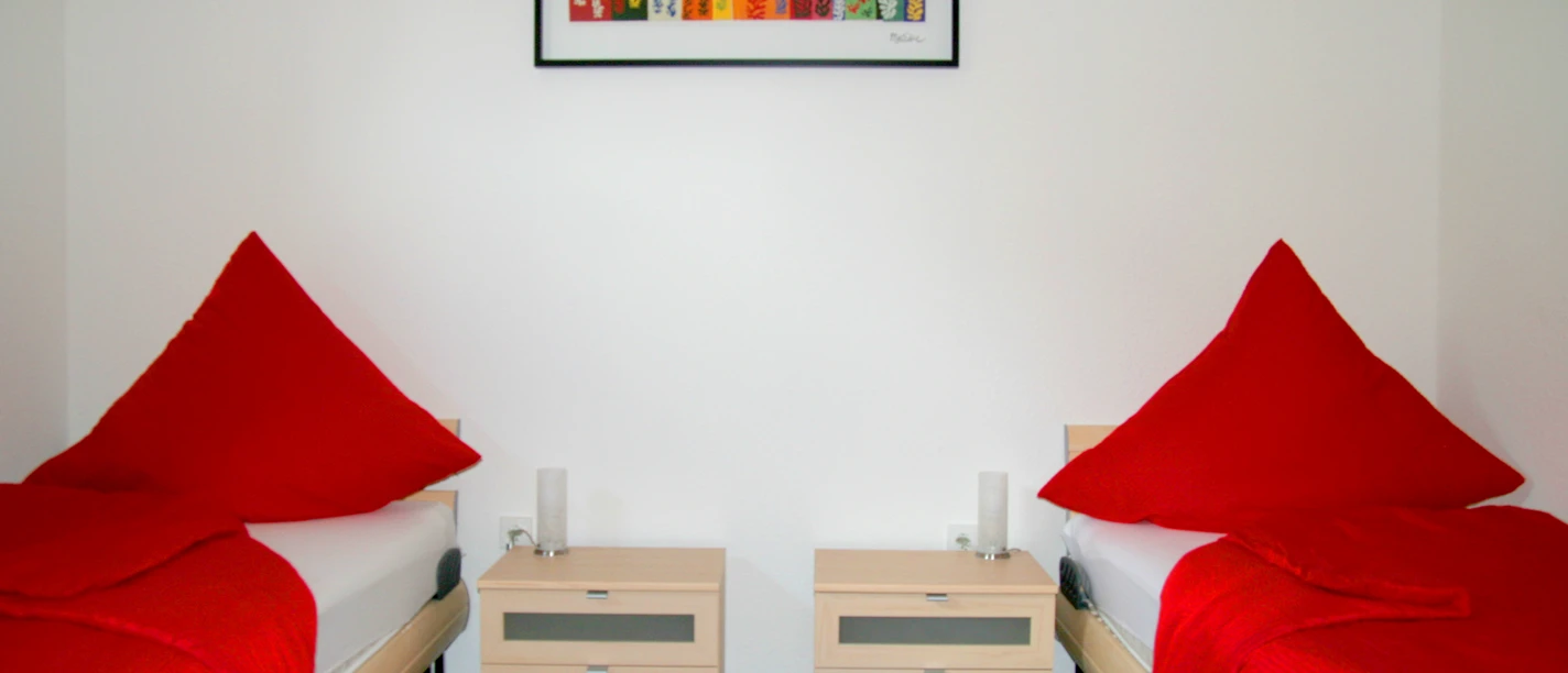 appartmentshaus_senne.jpg Zwei Einzelbetten mit roten Tagesdecken und zweckmäßigen Nachttischen in einem hellen Raum.
