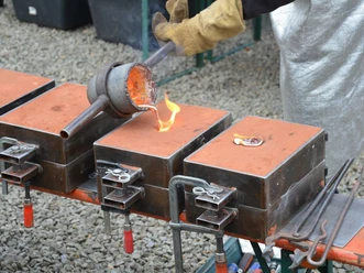 Metallgießen am Oelchenshammer Ein Handwerker gießt glühendes, flüssiges Metall in eine orangefarbene Sandform auf einem Tisch.