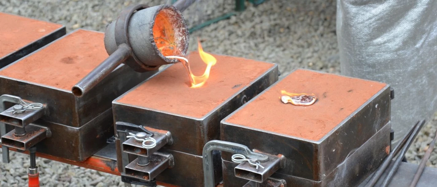 Metallgießen am Oelchenshammer Ein Handwerker gießt glühendes, flüssiges Metall in eine orangefarbene Sandform auf einem Tisch.