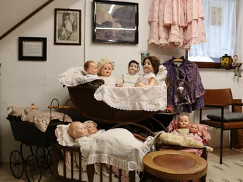 Spielzeugmuseum mit antiken Puppen in historischen Kinderwagen, umgeben von gerahmten Bildern.