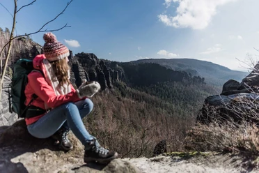 Uitzicht vanaf de "Heiligen Stiege" Een vrouw met een rode jas en pet zit op een rots bij het uitzichtpunt "Heilige Stiege" en kijkt uit over een bebost berglandschap onder een blauwe hemel.