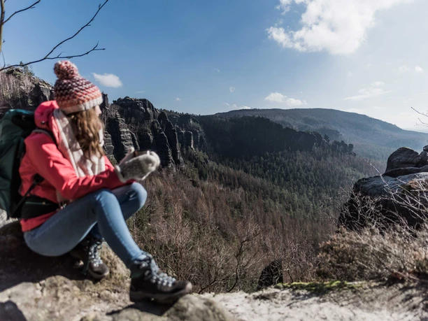Uitzicht vanaf de "Heiligen Stiege" Een vrouw met een rode jas en pet zit op een rots bij het uitzichtpunt "Heilige Stiege" en kijkt uit over een bebost berglandschap onder een blauwe hemel.
