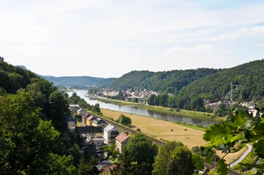 Carolahöhe in Krippen Blick auf eine Flusslandschaft mit grünen Hügeln, einem Dorf am Ufer und Feldern im Vordergrund unter einem klaren Himmel.