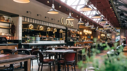 lo_osnabrueck_2_restaurantfoto_2.jpg Ein stilvolles Restaurant mit deckenhohen Fenstern, Holztischen, einem offenen Küchenbereich und warmen Licht.