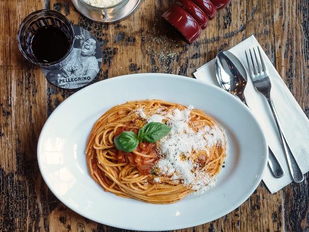 lo_osnabrueck_foodbild_pasta.jpg Spaghetti mit Tomatensauce, bestreut mit Parmesan und Basilikum auf Holztisch, daneben Rotweinglas.