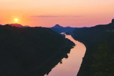 Klein bastion uitzicht Schmilka Zonsondergang over een rivier die door een heuvelachtig landschap kronkelt, met een oranjekleurige lucht en donkere silhouetten van de heuvels.