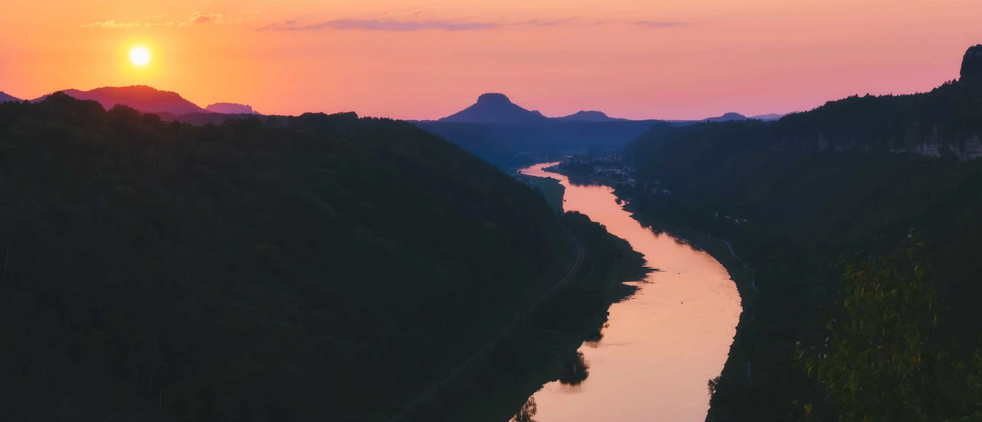 Kleine Bastei Aussicht Schmilka Sonnenuntergang über einem Fluss, der sich durch eine hügelige Landschaft schlängelt, mit einem orangefarbenen Himmel und dunklen Silhouetten der Hügel.