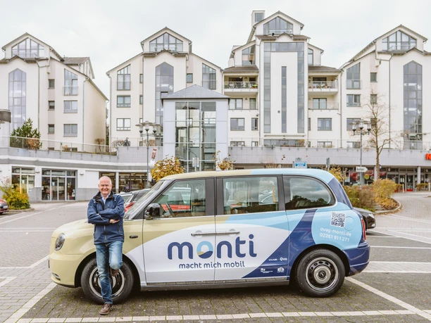 monti Ein Mann lehnt lächelnd an einem Elektro-Taxi, mit modernen Wohngebäuden im Hintergrund.