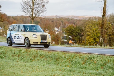 monti Taxi mit dem Schriftzug "mobi" fährt durch eine herbstliche Landschaft mit Hügeln und Wolken.
