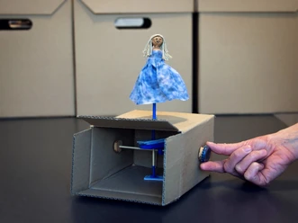 Automatenbox Eine blau gekleidete Puppe auf Kartonkonstruktion wird mit Hand an einer Kurbel bewegt.