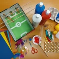 Angespielt Bastelmaterialien auf Tisch: Karton-Fußballspiel, Farben, Glitzerpapier, Scheren, Federn, und weitere Utensilien.