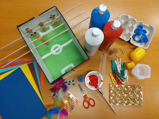 Angespielt Bastelmaterialien auf Tisch: Karton-Fußballspiel, Farben, Glitzerpapier, Scheren, Federn, und weitere Utensilien.