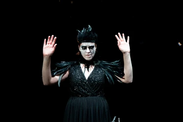 Theater im Bauturm A woman in a black dress with feather details wears dramatic make-up and raises her hands.
