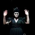Theater im Bauturm Eine Frau in schwarzem Kleid mit Federdetails trägt dramatisches Make-up und hebt die Hände.A woman in a black dress with feather details wears dramatic make-up and raises her hands.