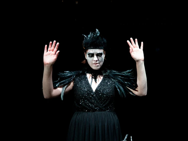 Theater im Bauturm A woman in a black dress with feather details wears dramatic make-up and raises her hands.