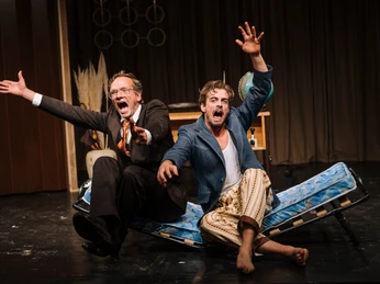 Theater im Bauturm Zwei Schauspieler in dramatischer Pose auf Bettenrahmen. Einer im Anzug, der andere leger. Two actors in dramatic poses on bed frames. One in a suit, the other casual.