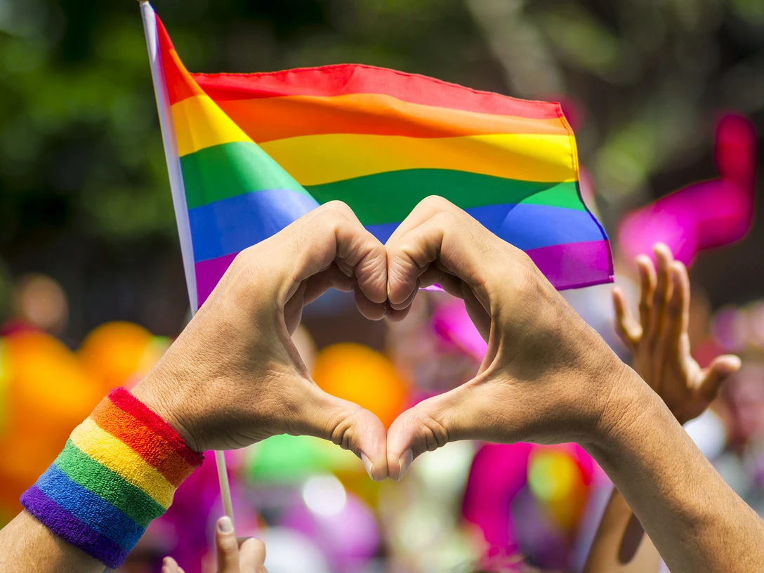 LGBTQ+-Herz Hände formen ein Herz vor einer RegenbogenflaggeHands form a heart in front of a rainbow flag