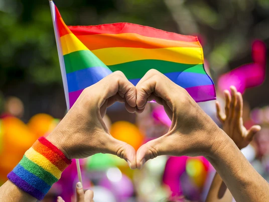 LGBTQ+-Herz Hände formen ein Herz vor einer RegenbogenflaggeHands form a heart in front of a rainbow flag